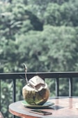 A fresh coconut with a straw and a spoon placed on a small plate sits on a wooden table. In the background, there is a lush, green forest creating a serene and refreshing atmosphere. A smartphone is also visible on the table next to the coconut.