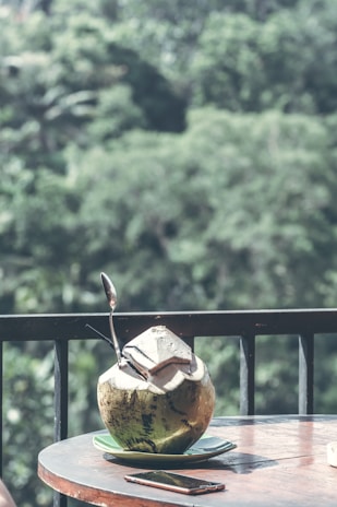 A fresh coconut with a straw and a spoon placed on a small plate sits on a wooden table. In the background, there is a lush, green forest creating a serene and refreshing atmosphere. A smartphone is also visible on the table next to the coconut.