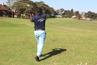 A person stands on a golf course, mid-swing, with a golf club in hand. Wearing a navy shirt and light blue pants, they are positioned on a patch of green grass. In the background, there are trees and several buildings with red roofs.