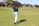 A person stands on a golf course, mid-swing, with a golf club in hand. Wearing a navy shirt and light blue pants, they are positioned on a patch of green grass. In the background, there are trees and several buildings with red roofs.