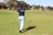 A person stands on a golf course, mid-swing, with a golf club in hand. Wearing a navy shirt and light blue pants, they are positioned on a patch of green grass. In the background, there are trees and several buildings with red roofs.