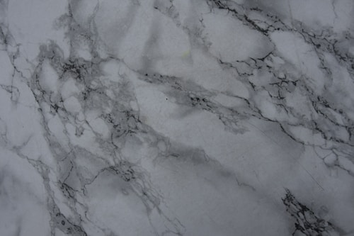 A close-up view of a marble surface featuring intricate gray veining patterns on a white background. The texture appears smooth and polished, with natural variations in the stone creating a sense of elegance and luxury.