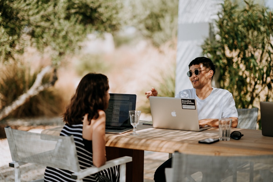 Two people are seated at a wooden outdoor table, each working on a laptop. The person on the right is wearing sunglasses and gesturing towards the person across the table, who is wearing a striped dress. The setting is bright and sunny, with greenery in the background. There are clear glasses and smartphones on the table.