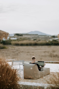 A person sits on a beige bean bag outdoors, working on a laptop. The setting appears to be a rural or natural landscape with dry grass and distant hills in the background. A white wooden chair is placed nearby. The scene is peaceful and open, with a subtle overcast sky.