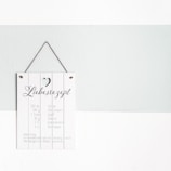 A decorative sign hangs on a light gray and white wall. The sign has a rustic wooden texture with a small heart cut-out at the top. It features playful typography with the word 'Liebesrezept' and a list of ingredients written below in German, suggesting a 'recipe for love.'