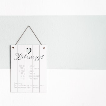 A decorative sign hangs on a light gray and white wall. The sign has a rustic wooden texture with a small heart cut-out at the top. It features playful typography with the word 'Liebesrezept' and a list of ingredients written below in German, suggesting a 'recipe for love.'