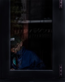 A person wearing a dark cap and glasses is partially visible through a window, seated with a contemplative expression. The dim lighting within the room contrasts with the brighter reflections on the glass, creating an introspective atmosphere.
