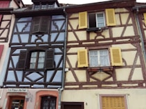 Two adjacent traditional half-timbered houses with vibrant colors. The house on the left features dark brown beams with pastel blue panels and dark shutters. The right house has brown beams and cream panels with bright yellow shutters. The building with the sign 'La Fleur de Sel' indicates a restaurant on the ground floor.