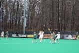 Several athletes are playing lacrosse on a field surrounded by tall, leafless trees. The players are actively engaged in the game, with some wearing green jerseys and others in white. The field has a bright green turf, and there is a banner in the background with motivational slogans.