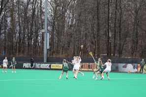 Several athletes are playing lacrosse on a field surrounded by tall, leafless trees. The players are actively engaged in the game, with some wearing green jerseys and others in white. The field has a bright green turf, and there is a banner in the background with motivational slogans.