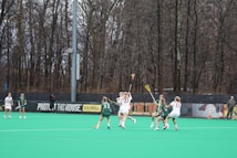 Several athletes are playing lacrosse on a field surrounded by tall, leafless trees. The players are actively engaged in the game, with some wearing green jerseys and others in white. The field has a bright green turf, and there is a banner in the background with motivational slogans.