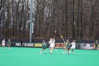 Several athletes are playing lacrosse on a field surrounded by tall, leafless trees. The players are actively engaged in the game, with some wearing green jerseys and others in white. The field has a bright green turf, and there is a banner in the background with motivational slogans.
