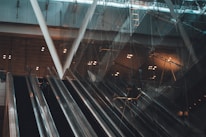 Several escalators ascend within a modern building with a glass facade. The lighting is dim, casting reflections and shadows across the surfaces. Structural beams and lighting fixtures contribute to the architectural design.