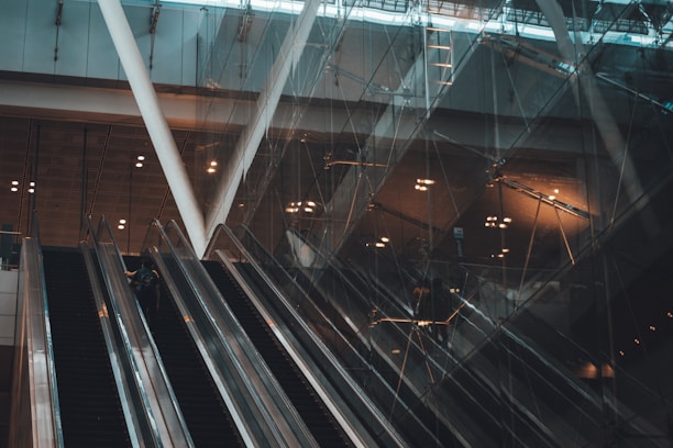 Several escalators ascend within a modern building with a glass facade. The lighting is dim, casting reflections and shadows across the surfaces. Structural beams and lighting fixtures contribute to the architectural design.
