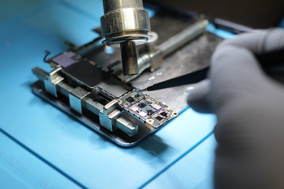 A close-up view of a person in gloves working on electronic circuitry with tools. The scene involves delicate handling of components on a circuit board, possibly for repair or assembly. A soldering tool is positioned near the board, indicating a process of adding or fixing components.