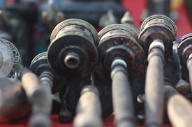 A detailed view of several intricately designed prayer wheels, mounted horizontally, with blurred handles extending towards the viewer. The objects have ornate engravings and a weathered appearance, indicating age and cultural significance. The background is softly out of focus, emphasizing the craftsmanship of the prayer wheels.