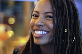A person with long braided hair and a nose piercing smiles warmly at the camera. The hair has decorative beads, and the background contains blurred lights creating a bokeh effect.