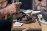 A close-up of a hand holding a wooden scoop, adding dried tea leaves into a small, rustic teapot. Several ceramic teacups and bowls are arranged on a wooden tray. The natural light and outdoor setting add a warm, inviting atmosphere.