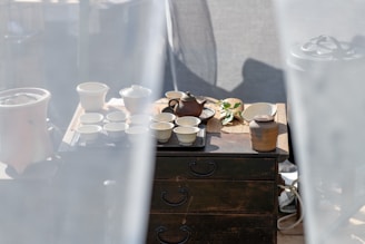 A rustic setup of a tea ceremony is arranged on a wooden table. There is a traditional teapot surrounded by multiple small ceramic cups and jars, with a few green leaves placed beside them. Soft white curtains frame the scene, creating a calm and serene ambiance.
