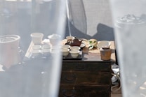 A rustic setup of a tea ceremony is arranged on a wooden table. There is a traditional teapot surrounded by multiple small ceramic cups and jars, with a few green leaves placed beside them. Soft white curtains frame the scene, creating a calm and serene ambiance.