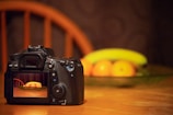 A digital camera rests on a wooden table, capturing an image of a blurred fruit arrangement in the background. The camera's screen displays a focused shot of a bowl containing oranges and a banana. The scene is softly lit, creating a warm and cozy atmosphere.