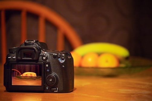 A digital camera rests on a wooden table, capturing an image of a blurred fruit arrangement in the background. The camera's screen displays a focused shot of a bowl containing oranges and a banana. The scene is softly lit, creating a warm and cozy atmosphere.