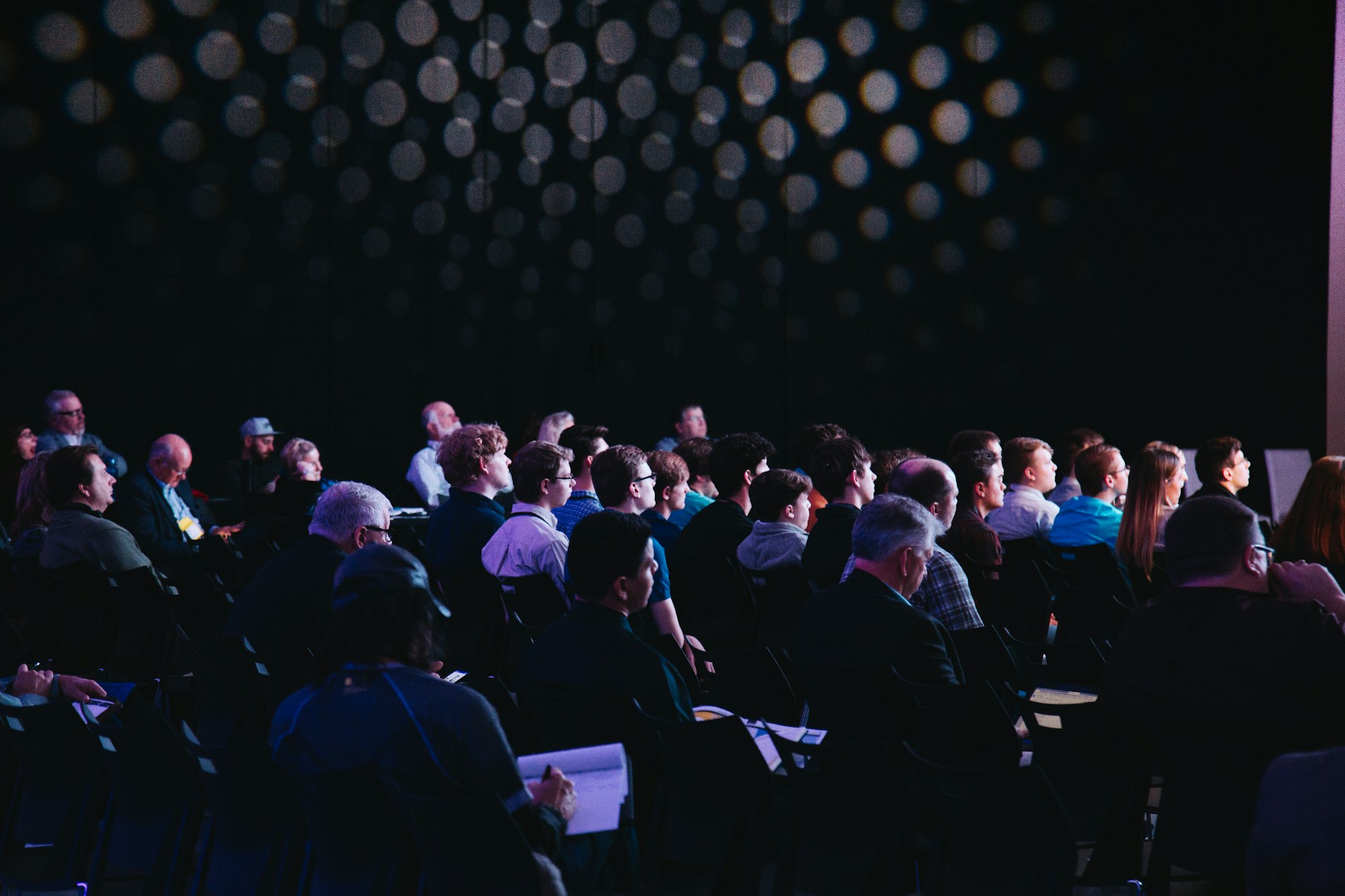 A group of people sitting down in a dark room, facing a stage