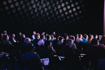A seated audience is attentively facing a stage or speaker, with a dark background featuring circular light patterns. The group consists of people of various ages, appearing focused on an unseen event or presentation.
