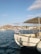 A small white boat docked in a marina with a Greek flag waving at the stern. The background features hills and a waterfront town with several other boats along the shoreline. The sun casts a shadow of a person waving on the side of the boat.