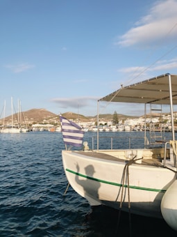 A small white boat docked in a marina with a Greek flag waving at the stern. The background features hills and a waterfront town with several other boats along the shoreline. The sun casts a shadow of a person waving on the side of the boat.