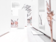 A minimalist architectural model featuring clean, white structures and walls interspersed with trees and plants that suggest an indoor garden or atrium. The overall environment is stark with a smooth wooden flooring and sparse foliage decoratively placed throughout.
