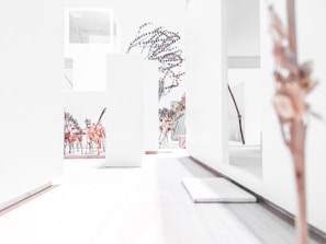A minimalist architectural model featuring clean, white structures and walls interspersed with trees and plants that suggest an indoor garden or atrium. The overall environment is stark with a smooth wooden flooring and sparse foliage decoratively placed throughout.