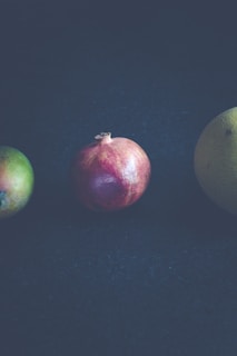 Three fruits placed in a row against a dark background. On the left, a green and slightly yellow mango. In the center, a red pomegranate. On the right, a melon with a rough, greenish texture.