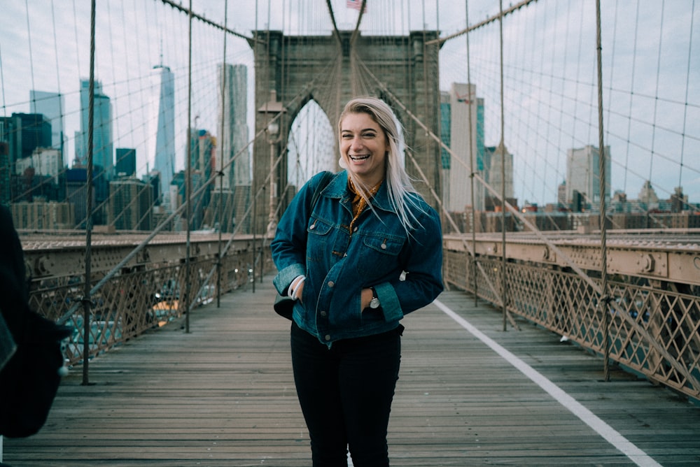 standing smiling woman wearing blue denim button-up jacket at Brooklyn Bridge