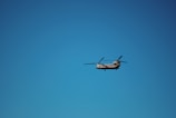 A helicopter with two rotors is flying against a clear blue sky. The aircraft is positioned in the center of the image, with its rotor blades visible in motion.