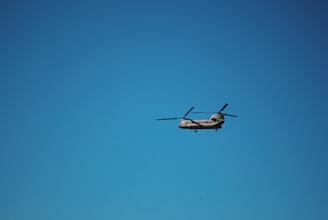 A helicopter with two rotors is flying against a clear blue sky. The aircraft is positioned in the center of the image, with its rotor blades visible in motion.
