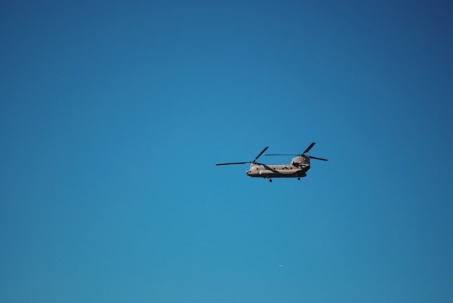 A helicopter with two rotors is flying against a clear blue sky. The aircraft is positioned in the center of the image, with its rotor blades visible in motion.