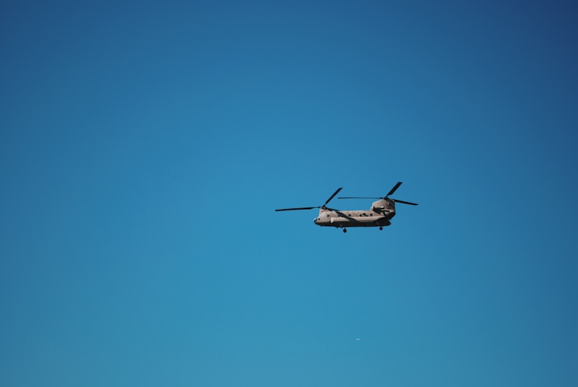 A helicopter with two rotors is flying against a clear blue sky. The aircraft is positioned in the center of the image, with its rotor blades visible in motion.