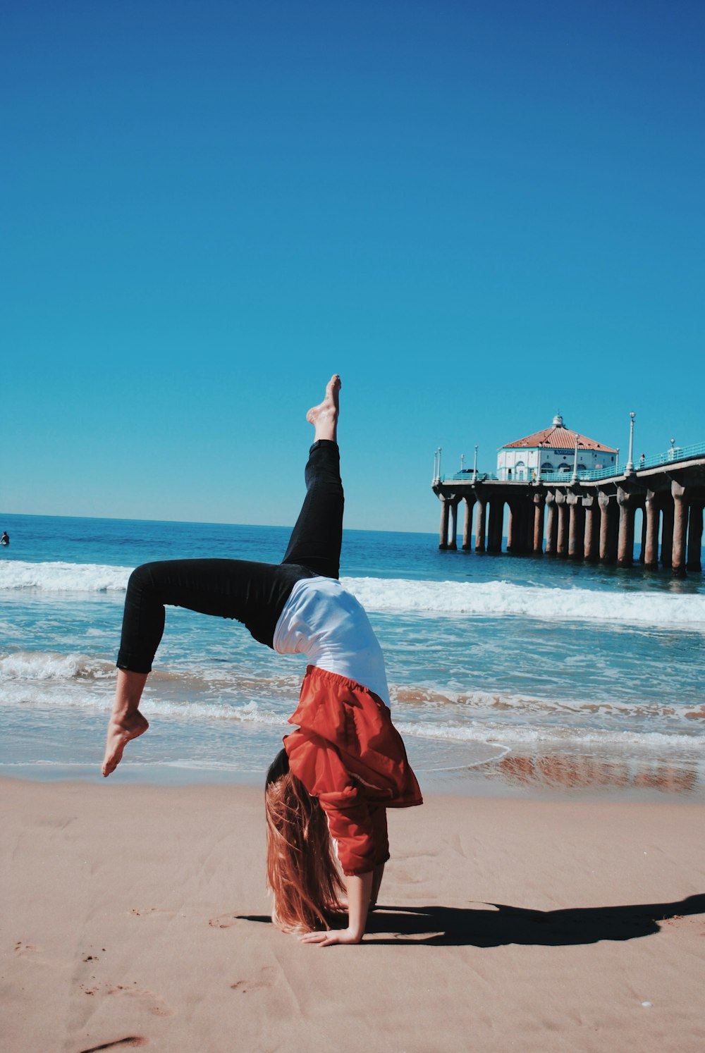 Frau mit roter Jacke und schwarzen Leggings macht Handstand an der Küste