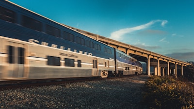 A high-speed passenger train is seen traveling along a railway track adjacent to a concrete bridge under a clear blue sky. The train has a sleek design and is moving rapidly, creating a sense of motion. The scene includes a mix of natural landscape with flora visible in the foreground.