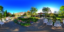A picturesque garden scene featuring a well-manicured lawn, colorful flower beds, and white metal chairs arranged along a cobblestone path. Tall trees and a historic building with red rooftops line the background under a vivid blue sky. The sun casts a warm glow, creating elongated shadows and an inviting atmosphere.