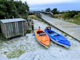 Two kayaks, one orange and one blue, are positioned on sandy ground near a wooden boardwalk surrounded by greenery. A sign near the kayaks indicates a kayak hand launch area. A small wooden structure, possibly for storage, is also nearby. The atmosphere is natural and serene, with a cloudy sky above.