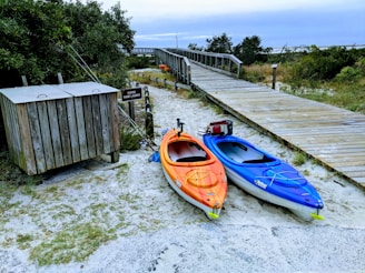 Two kayaks, one orange and one blue, are positioned on sandy ground near a wooden boardwalk surrounded by greenery. A sign near the kayaks indicates a kayak hand launch area. A small wooden structure, possibly for storage, is also nearby. The atmosphere is natural and serene, with a cloudy sky above.
