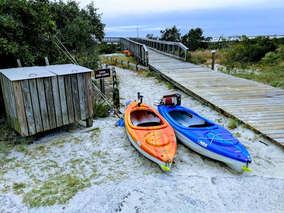 Two kayaks, one orange and one blue, are positioned on sandy ground near a wooden boardwalk surrounded by greenery. A sign near the kayaks indicates a kayak hand launch area. A small wooden structure, possibly for storage, is also nearby. The atmosphere is natural and serene, with a cloudy sky above.