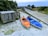 Two kayaks, one orange and one blue, are positioned on sandy ground near a wooden boardwalk surrounded by greenery. A sign near the kayaks indicates a kayak hand launch area. A small wooden structure, possibly for storage, is also nearby. The atmosphere is natural and serene, with a cloudy sky above.
