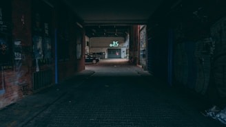 A dark alleyway leading to a partially lit open space with graffiti-covered walls and a distant car parked near a garage. Neon lights and posters add a mysterious, urban ambiance.