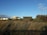 Several modern beach houses sit against a clear blue sky, framed by lush green bushes and dry, windswept grass in the foreground. The scene captures a tranquil seaside setting.