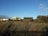 Several modern beach houses sit against a clear blue sky, framed by lush green bushes and dry, windswept grass in the foreground. The scene captures a tranquil seaside setting.