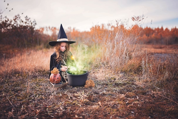 A young girl wearing a witch costume and hat is kneeling on the ground in a field. She is stirring a cauldron emitting a green, glowing mist. Nearby is a pumpkin lantern and dry, autumnal plant life surrounds the scene.