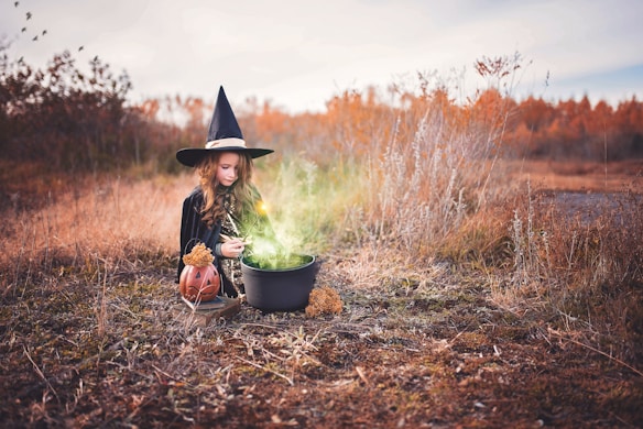 A young girl wearing a witch costume and hat is kneeling on the ground in a field. She is stirring a cauldron emitting a green, glowing mist. Nearby is a pumpkin lantern and dry, autumnal plant life surrounds the scene.